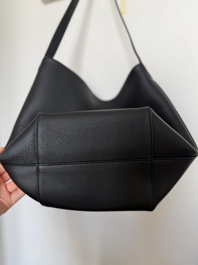 以下が含まれることがあります： 黒い革のショルダーバッグ。幅広のオープントップとシングルショルダーストラップが付いています。バッグは質感のある表面とシンプルでミニマリストなデザインで、日常使いに適しています。