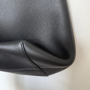 以下が含まれることがあります： 黒い革のトートバッグのクローズアップ。バッグはテクスチャ加工された表面と丸みを帯びた底の角を持っています。縫い目は縫い目に沿って見え、バッグの構造を強調しています。バッグのデザインはシンプルでエレガントです。