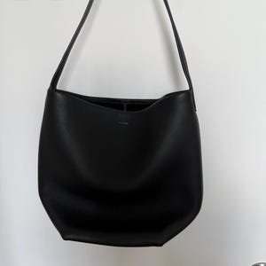 以下が含まれることがあります： 黒い革のショルダーバッグ。長いストラップが付いています。バッグはシンプルでミニマルなデザインで、広々としたインテリアです。白い壁に掛かっています。