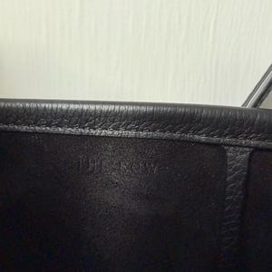 以下が含まれることがあります： 黒い革のハンドバッグのクローズアップ。バッグの上端が見え、テクスチャ加工された表面です。「THE ROW」のエンボス加工されたテキストがバッグの内側に配置されています。背景はプレーンなオフホワイトの壁です。