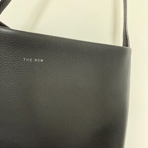 以下が含まれることがあります： 黒い革のショルダーバッグ。長いストラップが付いています。バッグは質感のある表面で、「THE ROW」のブランド名が銀色の文字で表示されています。