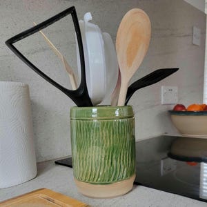 May include: A cylindrical green ceramic utensil holder with a textured vertical ribbed pattern and a smooth beige base. It contains a black triangular spatula, a white plastic mesh strainer, a wooden spoon, and a black slotted turner.