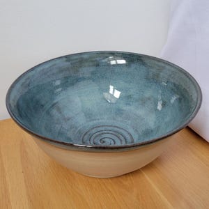 May include: A handmade ceramic bowl. The exterior is light tan, and the interior is a glossy teal. The bowl has a wide, shallow shape and a spiral design at the base. The rim is dark brown.