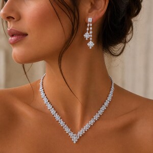 Può includere: Un set composto da collana e orecchini pendenti color argento, caratterizzato da un design a V decorato con numerose piccole pietre trasparenti e scintillanti. La collana presenta un motivo a chevron, mentre gli orecchini sono formati da due grappoli floreali pendenti.