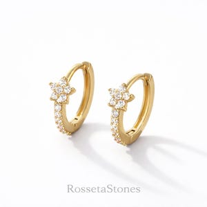 May include: A pair of gold hoop earrings featuring a star-shaped charm adorned with clear, sparkling stones. The front of each hoop is also embellished with a row of small, clear stones. The brand name RossetaStones is printed below the earrings.