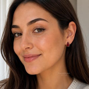 May include: A single teardrop-shaped earring featuring a smooth, deep red stone set in a simple metallic frame. The earring is worn on the earlobe, displaying a minimalist design with a polished finish and a vibrant, solid crimson colour.