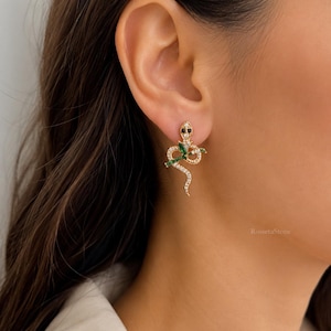 May include: A gold-toned snake earring featuring small clear pavé stones along the body, two dark stone eyes, and a green marquise-cut stone accent. The snake is coiled in a downward position with its tail curving at the bottom of the design.