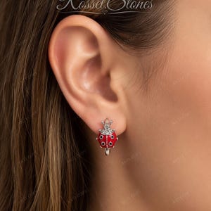 Pode incluir: Um brinco de argola em prata com um pingente de joaninha com corpo em esmalte vermelho brilhante, pontos pretos e um pequeno grupo de pedras transparentes no topo. O brinco possui um fecho articulado e exibe o texto Rosset Stones acima.