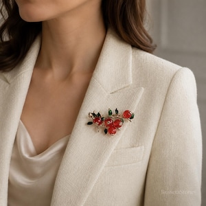 May include: A gold-toned brooch featuring three translucent red spherical berries and several small green leaves attached to a textured branch. The piece is pinned to the lapel of a cream-coloured blazer, showcasing a detailed botanical design.