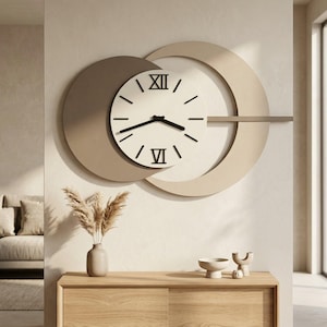 Op de afbeelding: Een moderne wandklok met een minimalistisch ontwerp. De klok heeft een witte wijzerplaat met zwarte Romeinse cijfers en wijzers, tegen een achtergrond van overlappende beige en taupe cirkels. De klok is bevestigd aan een neutraal gekleurde muur.