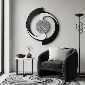 Puede incluir: Un reloj de pared moderno con esfera gris y detalles en negro, blanco y gris. El reloj está montado en una pared gris claro sobre un sillón negro con cojines decorativos. También se ven una mesa auxiliar y una lámpara de pie.