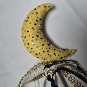 May include: A yellow felt crescent moon wand adorned with silver, gold, and black beads and star-shaped sequins. The wand features gold, black, and cream-colored ribbons and a wooden dowel handle. A whimsical, handmade item.