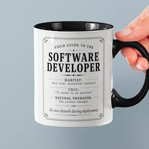 Könnte beinhalten: Weißer Keramikbecher mit schwarzem Henkel und Rand. Der Becher zeigt ein humorvolles "Field Guide to the Software Developer"-Design mit Text über seinen Lebensraum, seinen Ruf und seinen natürlichen Feind. Der Becher wird von einer Hand gehalten.