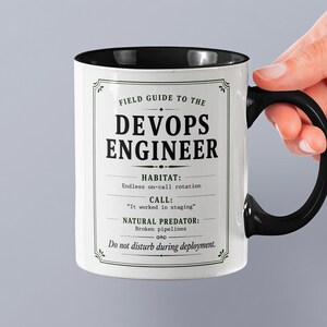 Puede incluir: Taza de cerámica blanca con interior y asa negros. La taza presenta el texto "FIELD GUIDE TO THE DEVOPS ENGINEER" con descripciones humorísticas del rol, incluyendo "HABITAT: Rotación de guardia constante."