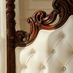 May include: Close-up of a luxurious bed headboard. The headboard features a tufted, off-white upholstered surface with crystal-like button accents. The frame is made of dark, intricately carved wood, showcasing ornate details and a classic design.