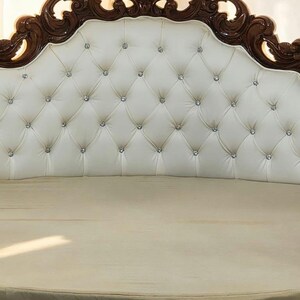 May include: A luxurious bed headboard with a tufted, off-white leather design. The headboard features a decorative, dark brown wooden frame with ornate carvings and embedded crystal accents. The bed frame is a light beige color.