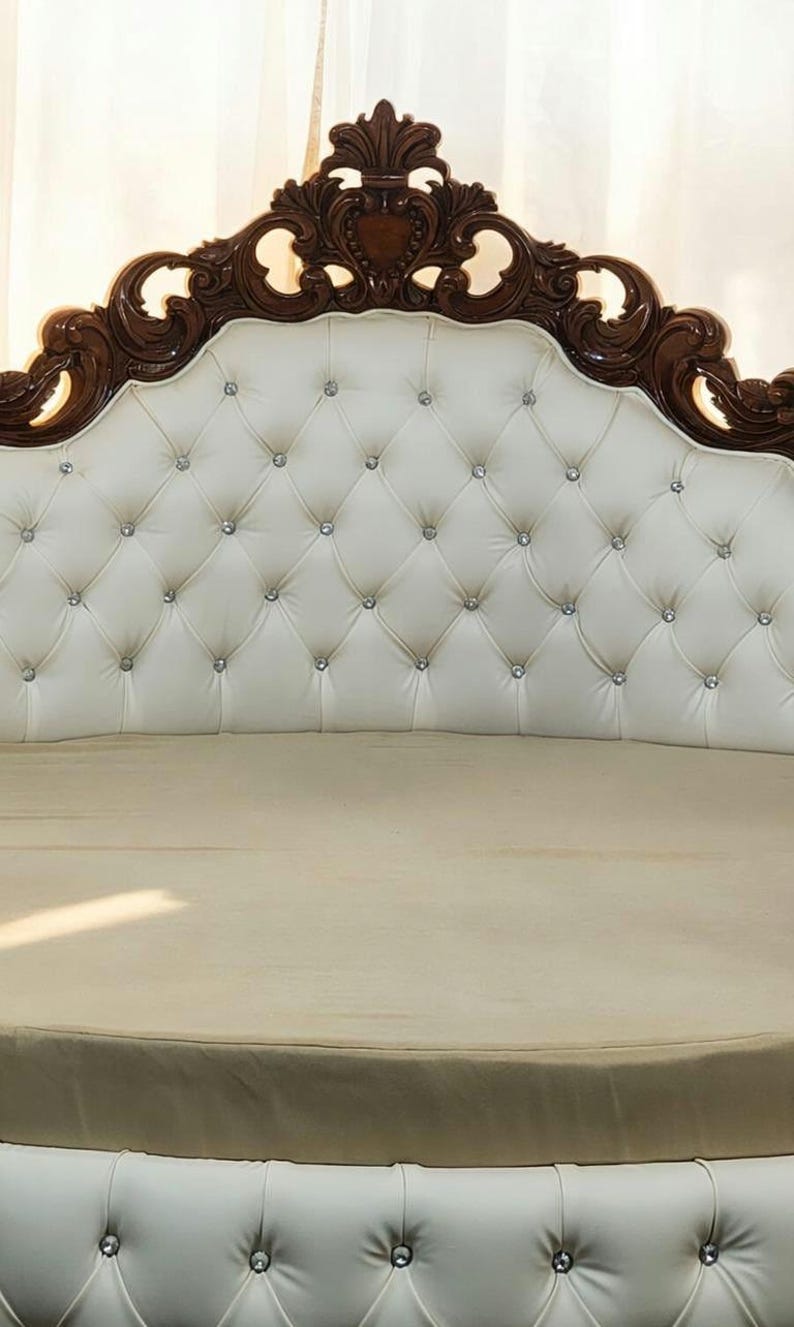 May include: A luxurious bed headboard with a tufted, off-white leather design. The headboard features a decorative, dark brown wooden frame with ornate carvings and embedded crystal accents. The bed frame is a light beige color.