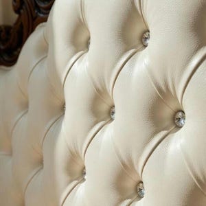 May include: Close-up of a cream-colored, tufted headboard with diamond-shaped crystal accents. The headboard has a dark brown, ornate wooden frame. The design is classic and elegant, suitable for a bedroom.