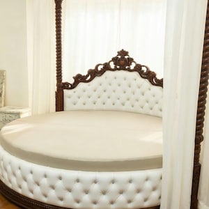 May include: A round bed with a white tufted headboard and base, accented with dark brown wood trim and posts. The bed has a cream-colored mattress and is draped with white curtains, creating a luxurious bedroom centerpiece.