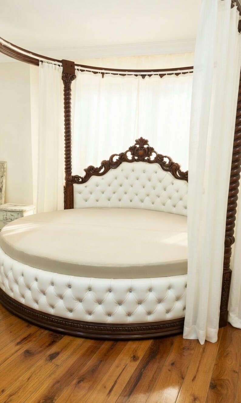 May include: A round bed with a white tufted headboard and base, accented with dark brown wood trim and posts. The bed has a cream-colored mattress and is draped with white curtains, creating a luxurious bedroom centerpiece.