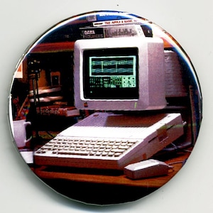 May include: A button featuring a vintage Apple computer with a keyboard, monitor, and mouse. The screen displays green text. The button has a white border and a background with books and other office supplies.