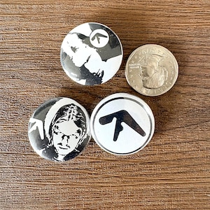 以下が含まれることがあります： 白黒のデザインが施された4つの丸いボタン。2つのボタンには肖像画、1つには様式化されたロゴ、銀貨が描かれています。ボタンは木製の表面に配置されています。