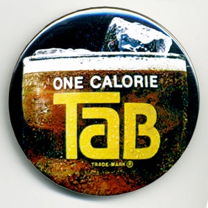 May include: A round, black and yellow button with a glass of brown soda with ice and the text "ONE CALORIE TAB TRADE-MARK" on it.