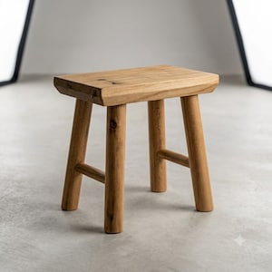 May include: A small, rectangular wooden stool featuring a natural light brown finish with visible wood grain and knots. The stool is constructed with four sturdy, cylindrical legs connected by horizontal support bars on the sides for stability.