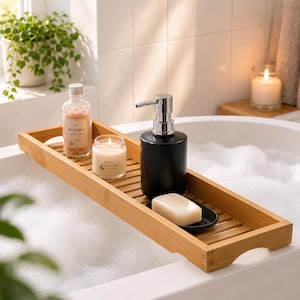 Könnte beinhalten: Ein rechteckiges Badewannenablage aus Bambus auf einer weißen, mit Schaum gefüllten Badewanne. Das Tablett enthält eine klare Glasflasche mit rosa Badesalz, eine kleine Kerze mit der Aufschrift Relax, einen schwarzen Seifenspender und ein Stück Seife.