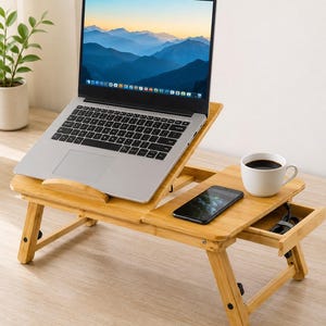 May include: A light brown bamboo laptop desk with an adjustable tilting surface, a side platform, and a small pull-out drawer. A silver laptop is open on the tilted section, while a black smartphone and a white ceramic mug sit on the flat side.