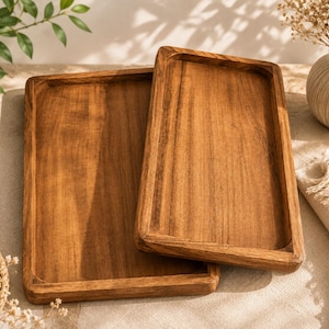 May include: Two rectangular wooden trays with a natural brown grain finish. The trays feature raised edges and a smooth, flat surface. One tray is positioned horizontally beneath the other, which is placed at a slight angle, showcasing the wood.