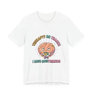 Puede incluir: Camiseta blanca con un diseño de cerebro de dibujos animados sosteniendo una taza de café. El texto "¿Terapia? No, gracias, tengo sobrepensamiento" rodea el cerebro.