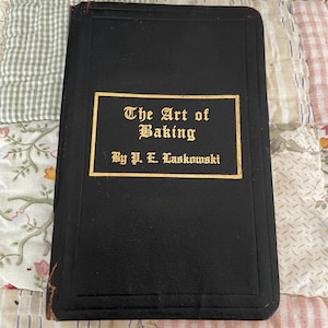 May include: A black hardcover book titled The Art of Baking by P. E. Laskowski. The front cover features a rectangular gold-bordered inset with gold lettering in a gothic-style font. The book has visible wear along the left spine edge.
