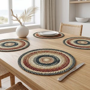 Puede incluir: Cuatro manteles individuales redondos trenzados en tonos rojos, beige y azules dispuestos sobre una mesa de madera clara. Un jarrón blanco con una planta seca y un cuchillo también están sobre la mesa. Los manteles individuales tienen un diseño en espiral.