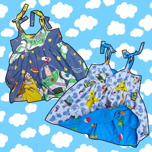 Könnte beinhalten: Zwei ärmellose Kleider mit Bindeträgern an den Schultern. Das linke Kleid ist dunkelblau mit einem Buzz Lightyear und Woody Aufdruck. Das rechte Kleid ist weiß mit einem bunten Pokemon-Muster und einem hellblauen Futter mit Pokebällen.