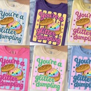 May include: Six t-shirts in different colours, each with a graphic design. The design features the text "You're a glitter dumpling" with a cartoon dumpling in a bamboo steamer. The shirts are in cream, purple, yellow, pink, tan, and blue.