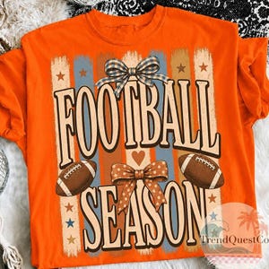 May include: An orange t-shirt featuring a graphic print with the text FOOTBALL SEASON in large brown letters. The design includes two brown footballs, a blue and white plaid bow, a brown polka dot bow, and vertical stripes with stars.