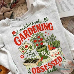 Puede incluir: Una sudadera gris claro con tem&aacute;tica de jardiner&iacute;a. El dise&ntilde;o incluye las palabras "members only GARDENING OBSESSED club" en rojo y verde, junto con ilustraciones de verduras, sobres de semillas y herramientas de jardiner&iacute;a.