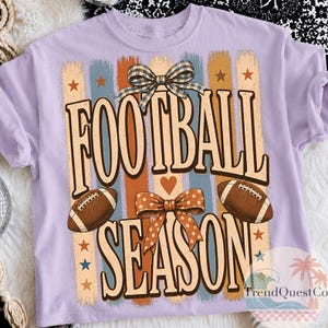 May include: A light purple t-shirt featuring a graphic print with the text FOOTBALL SEASON in large letters. The design includes two brown footballs, a blue and white checkered bow, an orange polka dot bow, and vertical stripes with stars.