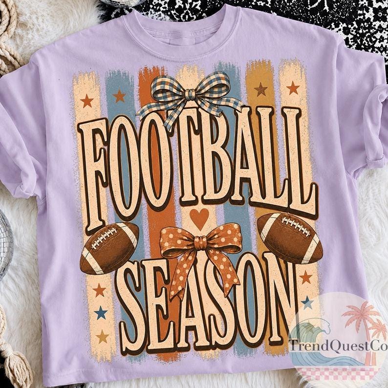 May include: A light purple t-shirt featuring a graphic print with the text FOOTBALL SEASON in large letters. The design includes two brown footballs, a blue and white checkered bow, an orange polka dot bow, and vertical stripes with stars.
