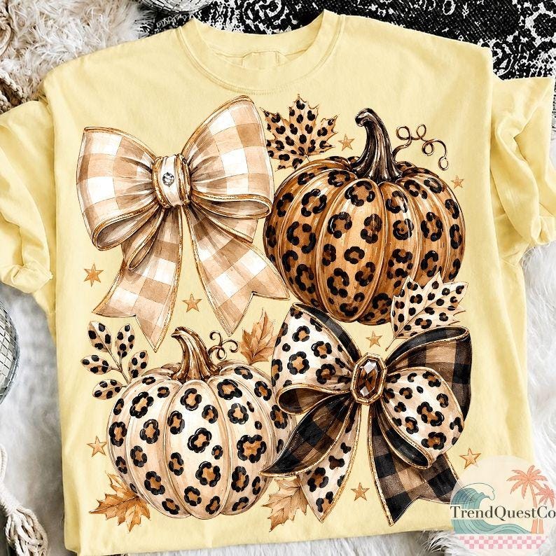 K&ouml;nnte beinhalten: Ein hellgelbes T-Shirt mit einem Grafikdruck von zwei K&uuml;rbissen mit Leopardenmuster, zwei gro&szlig;en dekorativen Schleifen in beigem und schwarzem Karomuster, verstreuten Herbstbl&auml;ttern und kleinen goldenen Sternen auf der Vorderseite.