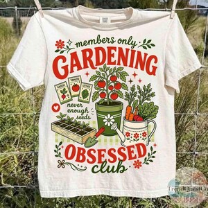 Puede incluir: Camiseta blanca con tem&aacute;tica de jardiner&iacute;a. La camiseta presenta el texto "members only GARDENING OBSESSED club" en rojo y verde, junto con ilustraciones de sobres de semillas, una planta de tomate, zanahorias y una regadera.