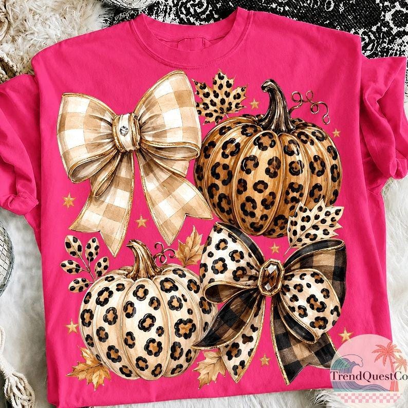 K&ouml;nnte beinhalten: Ein leuchtend pinkfarbenes T-Shirt mit einem Grafikdruck von zwei K&uuml;rbissen mit Leopardenmuster, zwei gro&szlig;en dekorativen Schleifen mit Karomuster und Leopardenmuster, verstreuten goldenen Sternen und Herbstbl&auml;ttern in Braun- und Beiget&ouml;nen.