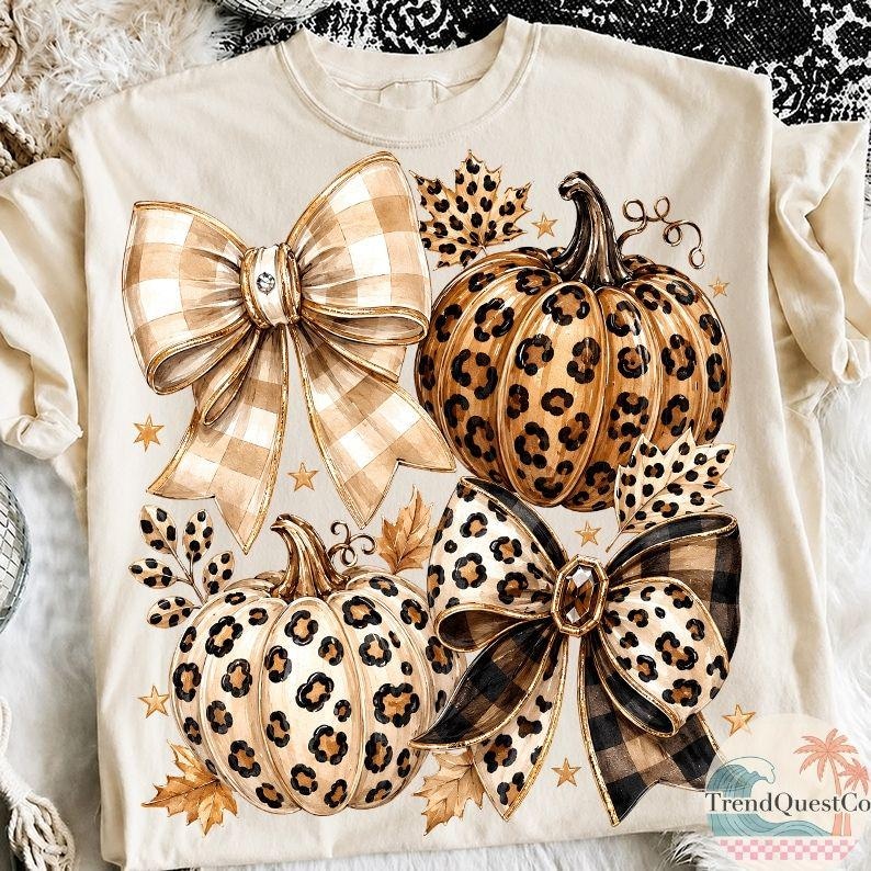 K&ouml;nnte beinhalten: Ein cremefarbenes T-Shirt mit einem Grafikdruck von zwei K&uuml;rbissen mit Leopardenmuster, zwei gro&szlig;en dekorativen Schleifen mit Karomuster und Leopardenprint sowie verstreuten Herbstbl&auml;ttern und kleinen Sternen auf der Vorderseite.