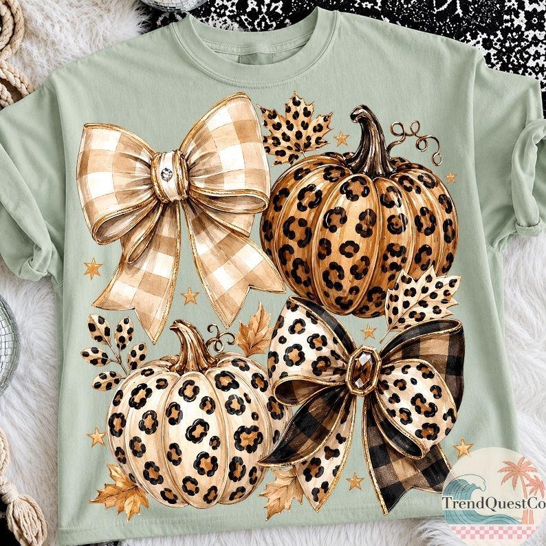 K&ouml;nnte beinhalten: Ein hellgr&uuml;nes T-Shirt mit einem Grafikdruck von zwei K&uuml;rbissen mit Leopardenmuster, zwei gro&szlig;en dekorativen Schleifen in beigem und schwarzem Karomuster, verstreuten Herbstbl&auml;ttern und kleinen goldenen Sternen. Der Text TrendQuestCo ist sichtbar.