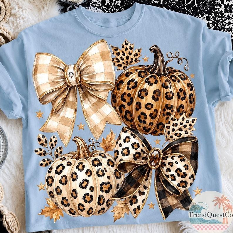 K&ouml;nnte beinhalten: Ein hellblaues T-Shirt mit einem Grafikdruck von zwei K&uuml;rbissen und zwei gro&szlig;en Schleifen. Die K&uuml;rbisse haben ein beiges und schwarzes Leopardenmuster. Die Schleifen zeigen ein beiges und wei&szlig;es Karomuster sowie ein beiges und schwarzes Leopardenmuster.
