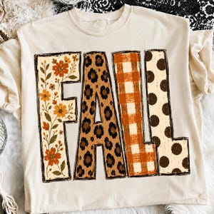 May include: A cream-colored t-shirt featuring the word FALL in large block letters. Each letter has a distinct pattern: the F has orange flowers and green leaves, the A has a brown leopard print, the L has an orange plaid, and the L has brown dots.