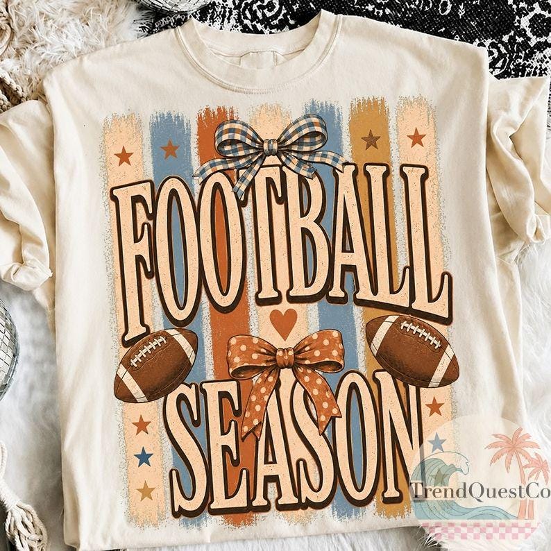May include: A cream-colored t-shirt featuring a graphic print with the text FOOTBALL SEASON in large brown letters. The design includes two brown footballs, a blue and white plaid bow, an orange polka dot bow, stars, and vertical stripes.