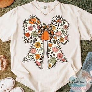 May include: A cream-coloured short-sleeve t-shirt featuring a large central graphic of a bow shape composed of orange pumpkins, pink and yellow flowers, green leaves, and leopard print patterns. The shirt has a crew neckline and a tag at the collar.