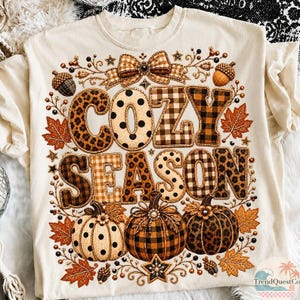 May include: A cream-colored t-shirt featuring a graphic print with the text COZY SEASON in large letters. The design includes pumpkins, autumn leaves, acorns, and a bow, decorated with leopard print, polka dots, and plaid patterns in brown tones.